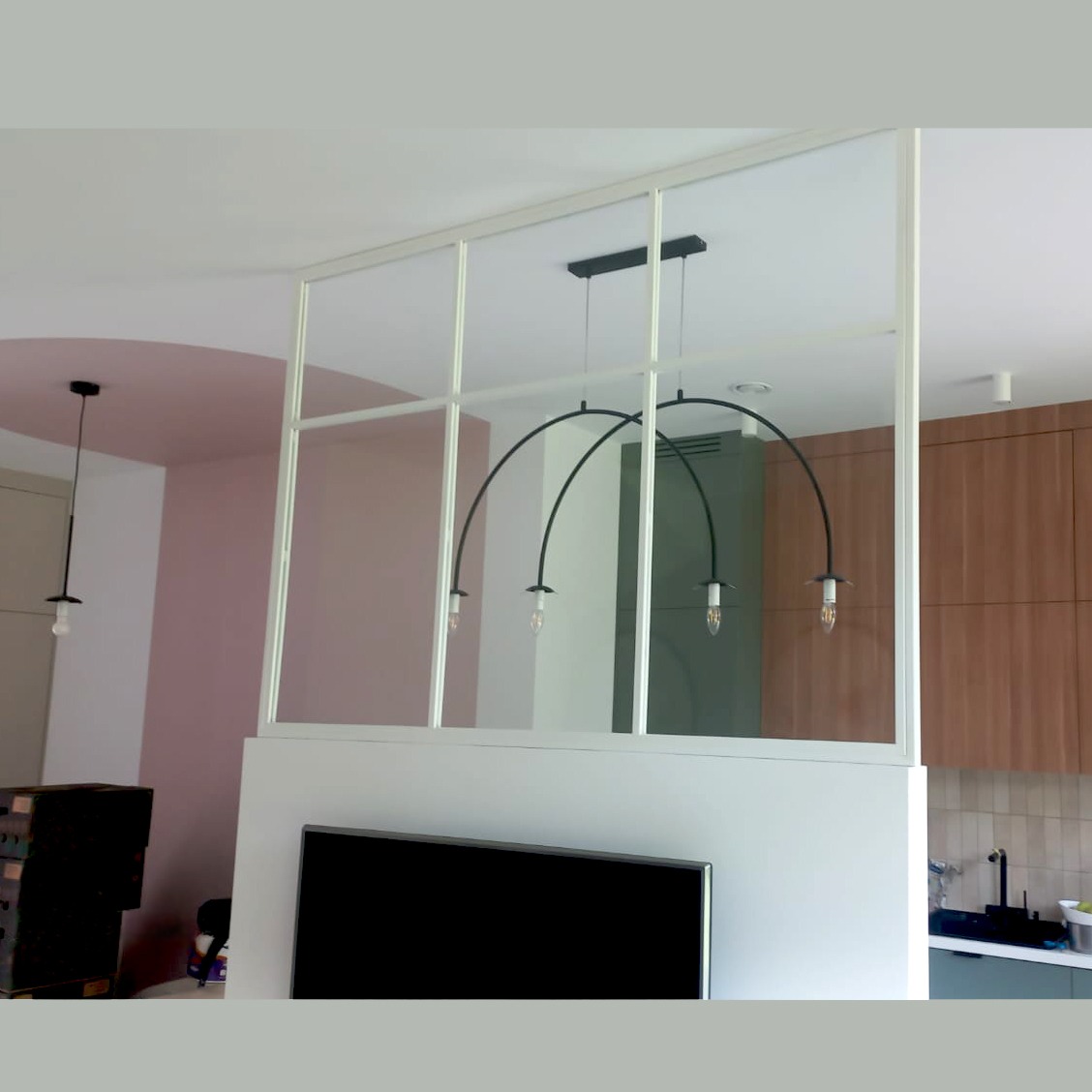 Nowoczesna aranżacja wnętrza z szklaną ścianką działową z białymi ramami, oddzielającą salon od kuchni, z widocznym telewizorem i designerskimi lampami.