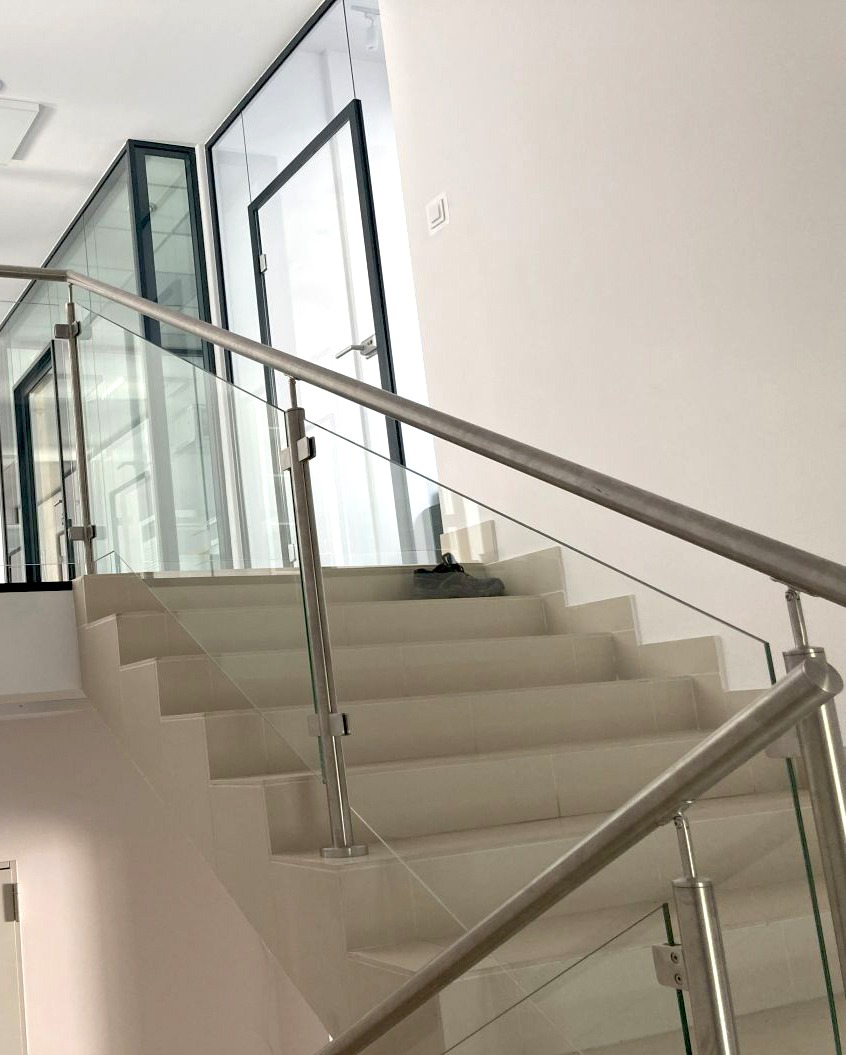 Schody wyłożone jasnymi płytkami z balustradą ze stali nierdzewnej i szkła, widoczne buty na schodach, w tle przeszklone drzwi z czarnymi ramami.