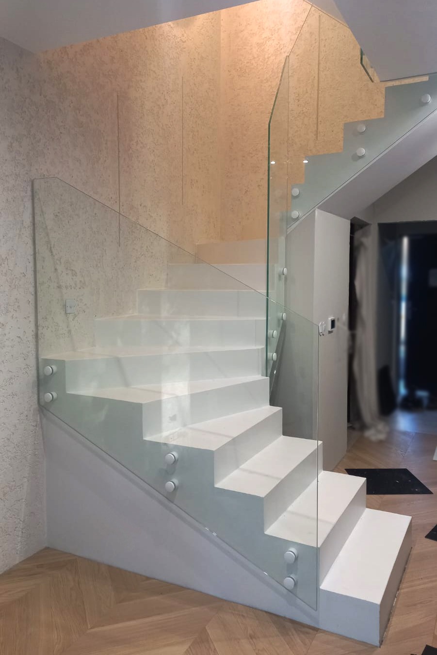 Nowoczesne, białe schody z przezroczystą szklaną balustradą mocowaną punktowo, widoczne w jasnym wnętrzu z drewnianą podłogą w jodełkę.