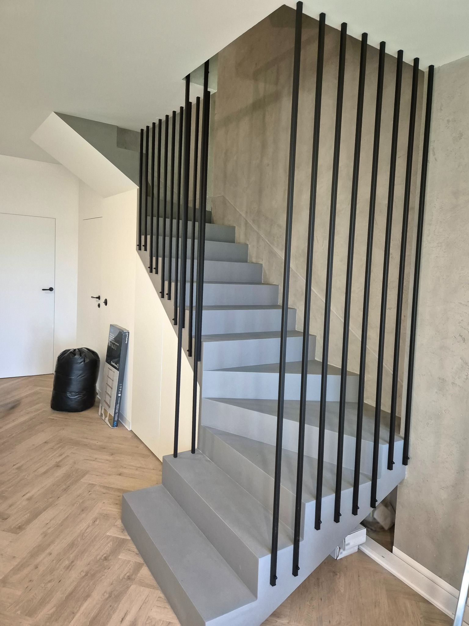 Nowoczesne schody z szarymi stopniami i minimalistyczną, czarną balustradą z pionowych prętów, na tle ściany o fakturze betonu i parkietu w jodełkę.