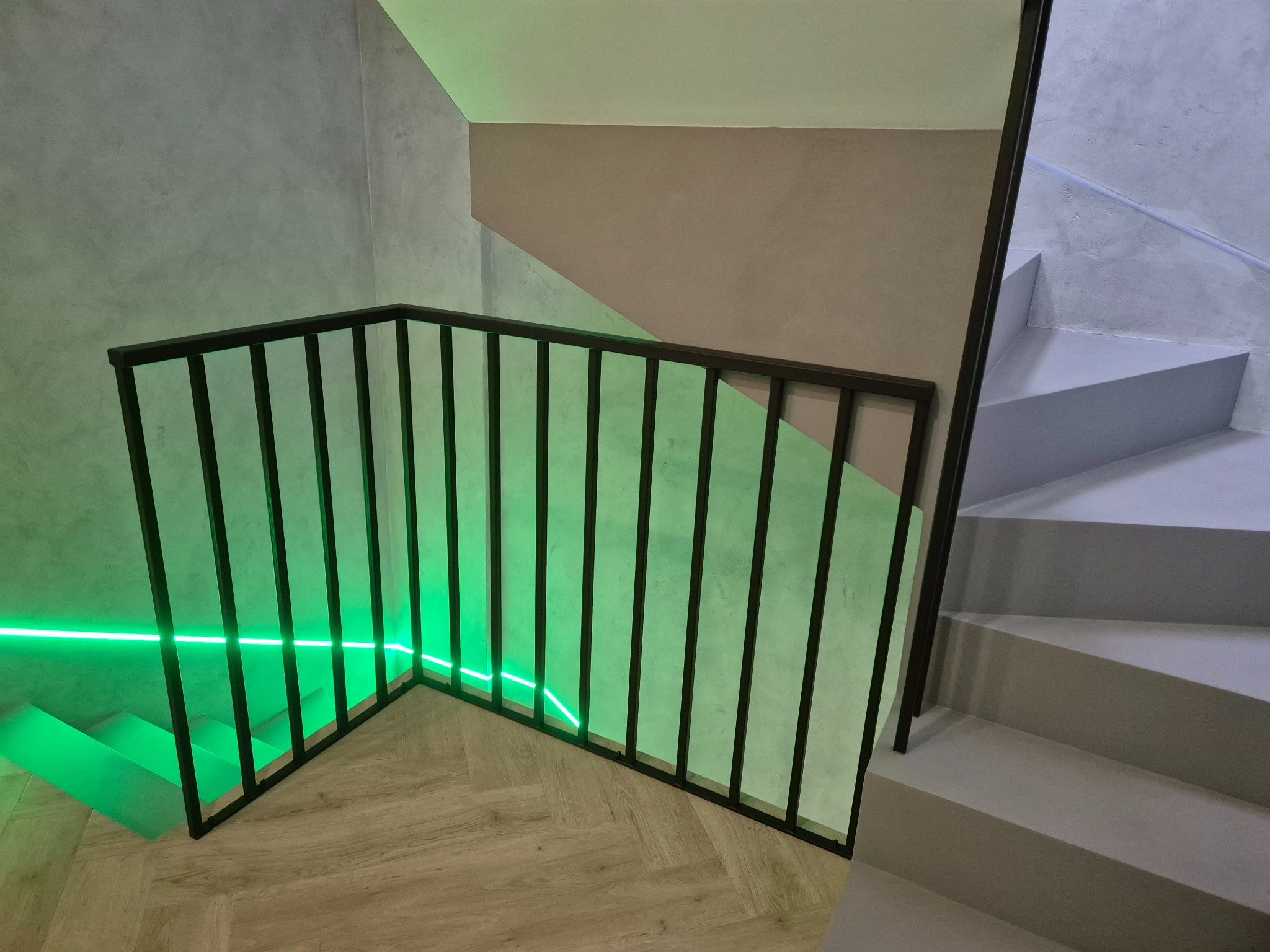 Schody z czarną, metalową balustradą podświetloną neonowym, zielonym światłem LED, widok z dołu na parkietową podłogę.