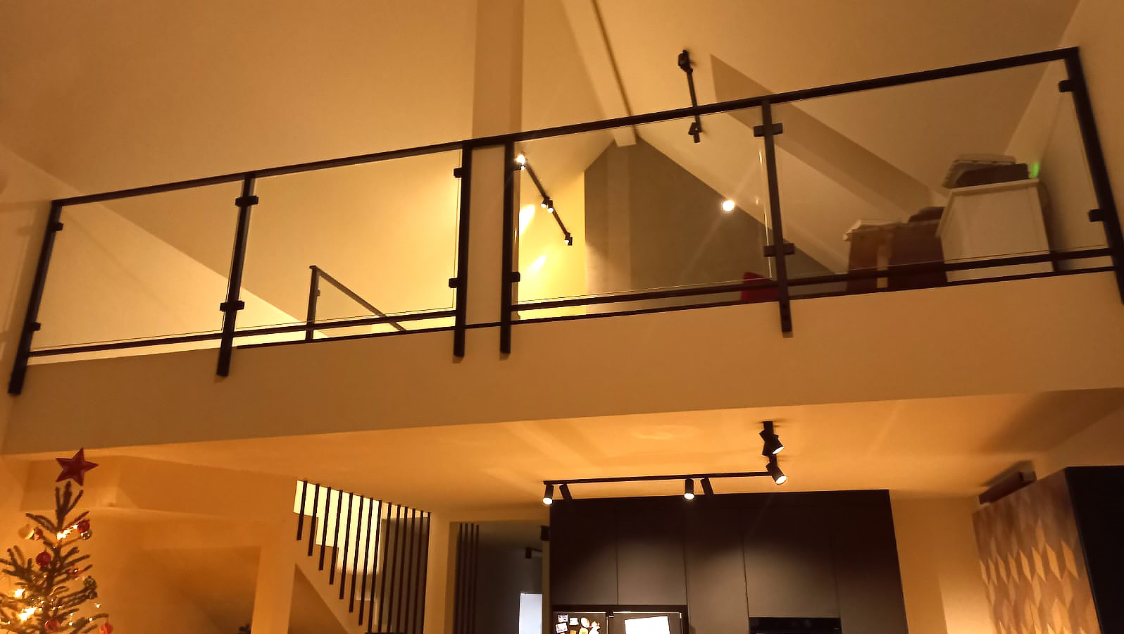 Wnętrze domu z antresolą zabezpieczoną szklaną balustradą z czarnymi, metalowymi słupkami, widok z dołu na antresolę, w tle schody i choinka.