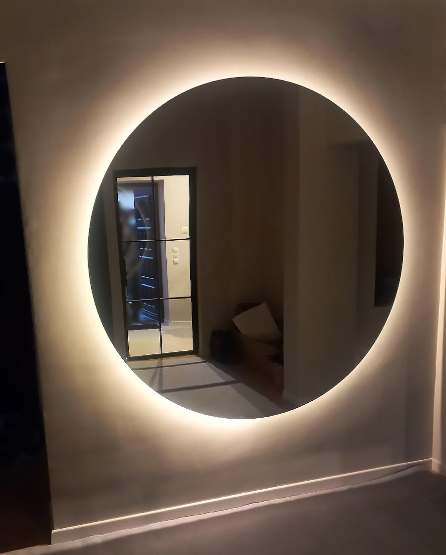 Okrągłe lustro z podświetleniem LED na ścianie, odbijające widok na czarne, szklane drzwi wewnętrzne.