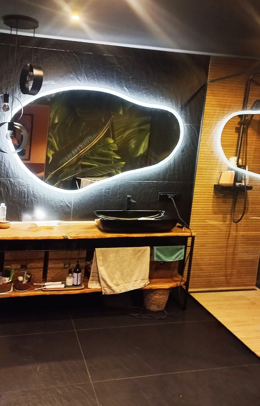 Nowoczesna łazienka z nieregularnym lustrem podświetlanym LED, drewnianym blatem pod umywalkę i czarną, owalną umywalką nablatową, uzupełniona o bambusowe akcenty i ciemną, kamienną okładzinę ściany.