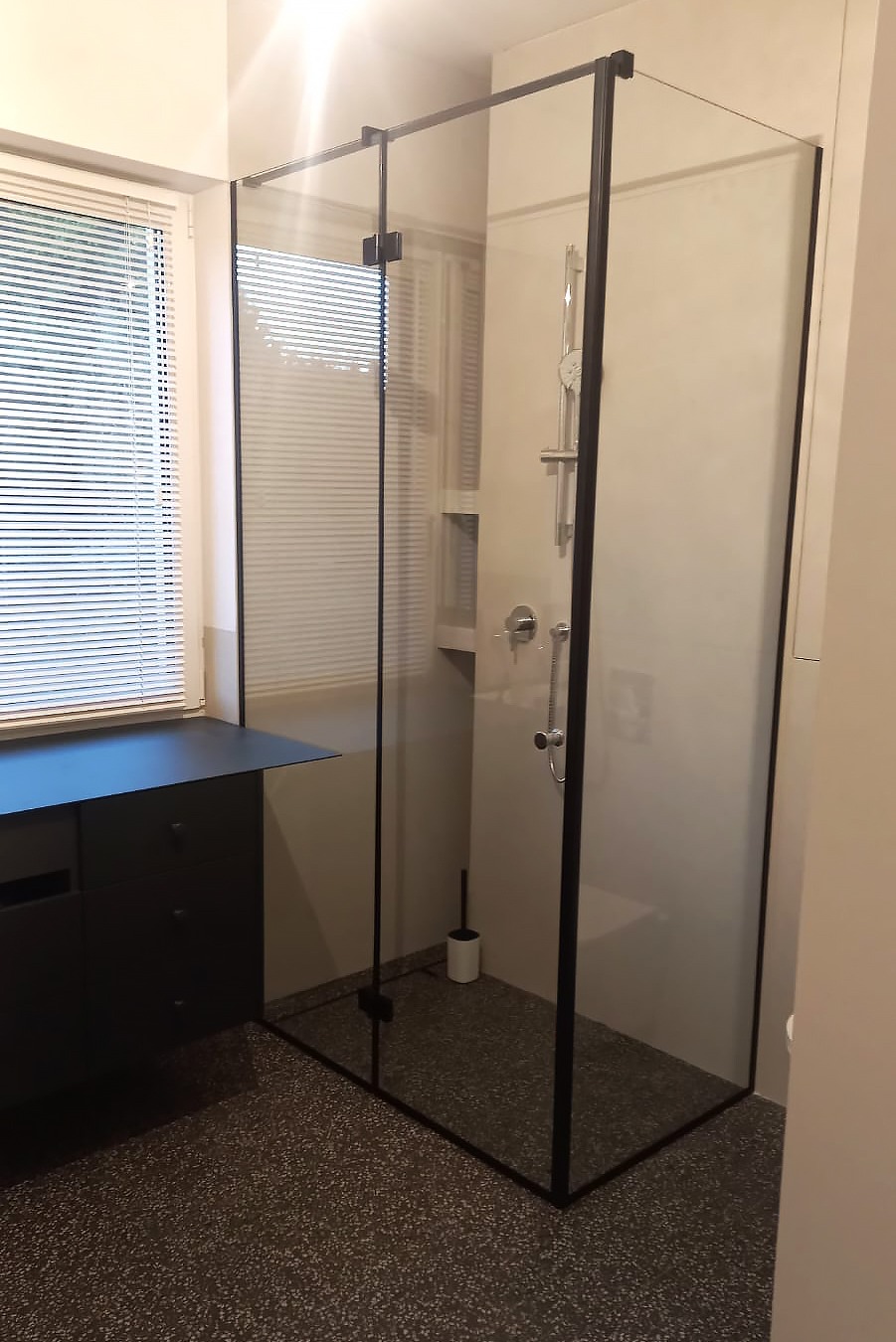 Nowoczesna kabina prysznicowa z czarnymi profilami i przezroczystym szkłem, widok z boku na tle okna z żaluzjami i czarnej szafki, na podłodze lastryko.
