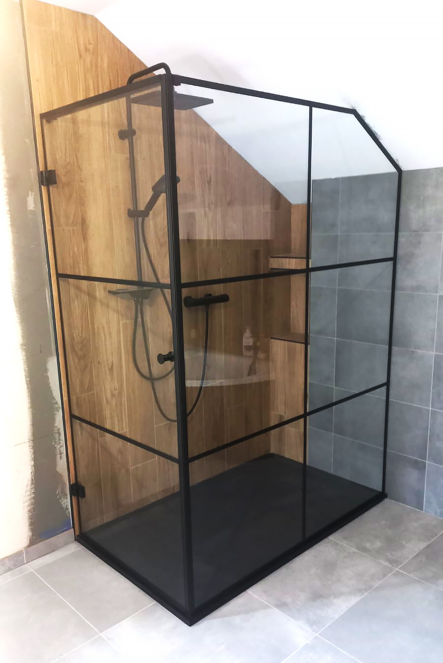 Kabina prysznicowa walk-in z czarnymi profilami i szybami, zamontowana w łazience z drewnopodobnymi i szarymi płytkami, z widoczną półką wewnątrz.