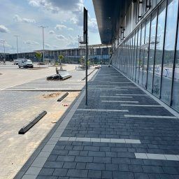 GEKKO SPÓŁKA Z OGRANICZONĄ ODPOWIEDZIALNOŚCIĄ - Nowa, grafitowa kostka brukowa z jasnymi wstawkami ułożona przy nowoczesnym budynku z przeszkloną fasadą. Widoczne elementy infrastruktury i pracownicy na rusztowaniu.