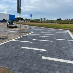 GEKKO SPÓŁKA Z OGRANICZONĄ ODPOWIEDZIALNOŚCIĄ - Nowo ułożona, ciemnoszara kostka brukowa na parkingu z białymi liniami wyznaczającymi miejsca postojowe. W tle widoczny budynek i sprzęt budowlany.