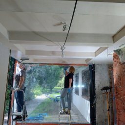 GEKKO SPÓŁKA Z OGRANICZONĄ ODPOWIEDZIALNOŚCIĄ - Dwie osoby na drabinach szpachlują sufit w pomieszczeniu z dużym oknem. Widoczne narzędzia, zabezpieczenia i fragmenty surowej ściany z cegły.
