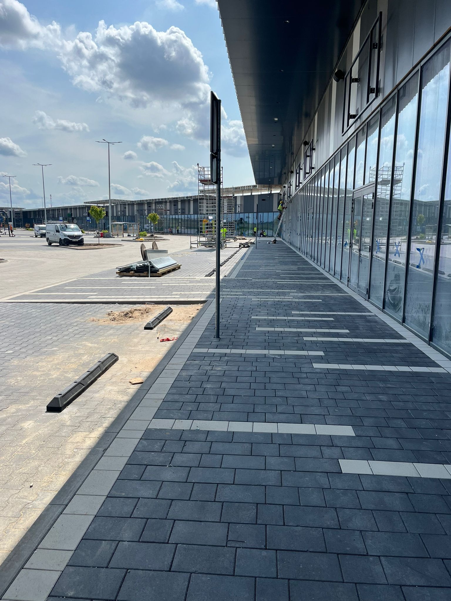 Nowa, grafitowa kostka brukowa z jasnymi wstawkami ułożona przy nowoczesnym budynku z przeszkloną fasadą. Widoczne elementy infrastruktury i pracownicy na rusztowaniu.