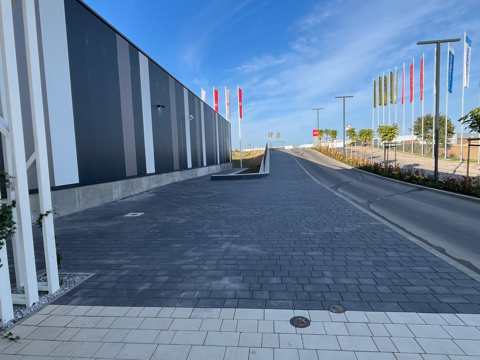Nowoczesna nawierzchnia z kostki brukowej w odcieniach szarości, prowadząca wzdłuż budynku z panelami w paski, z flagami w tle i elementami zieleni.