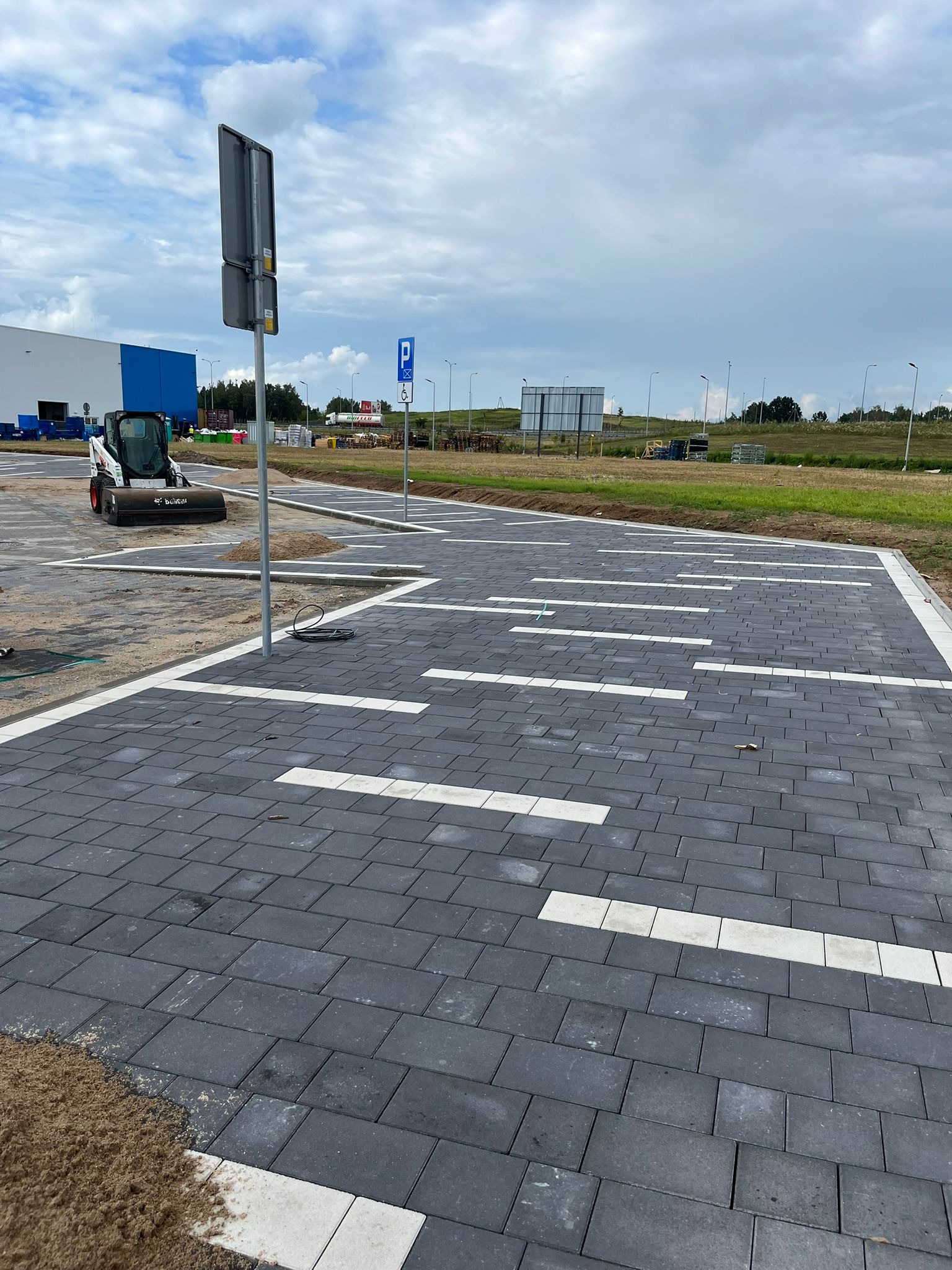 Nowo ułożona, ciemnoszara kostka brukowa na parkingu z białymi liniami wyznaczającymi miejsca postojowe. W tle widoczny budynek i sprzęt budowlany.