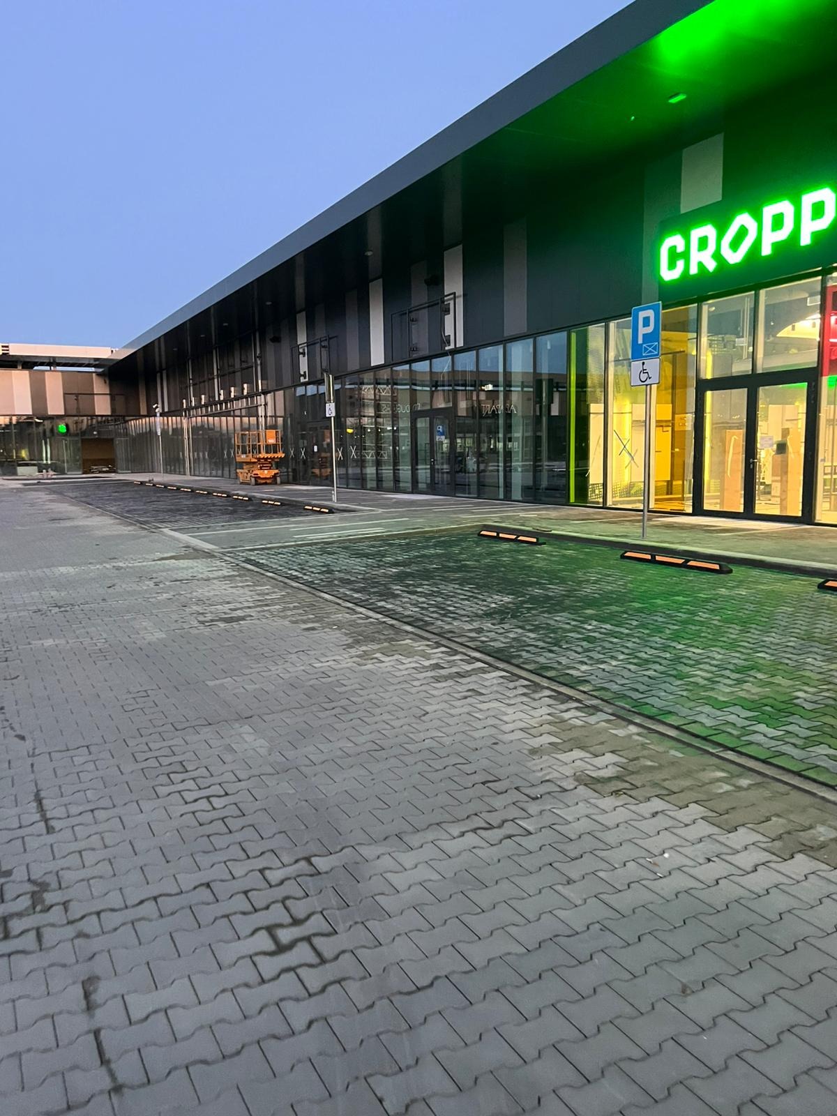 Szara kostka brukowa przed budynkiem sklepu Cropp z zielonymi akcentami. Widoczny znak parkingu dla niepełnosprawnych i podnośnik koszowy w tle.