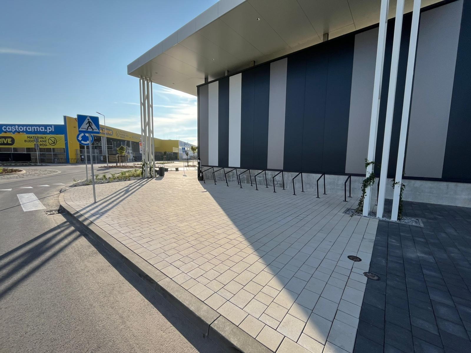 Jasnoszara kostka brukowa przed budynkiem z czarno-szarymi panelami. W tle widoczny sklep budowlany Castorama i błękitne niebo. Cień rzucany przez budynek.