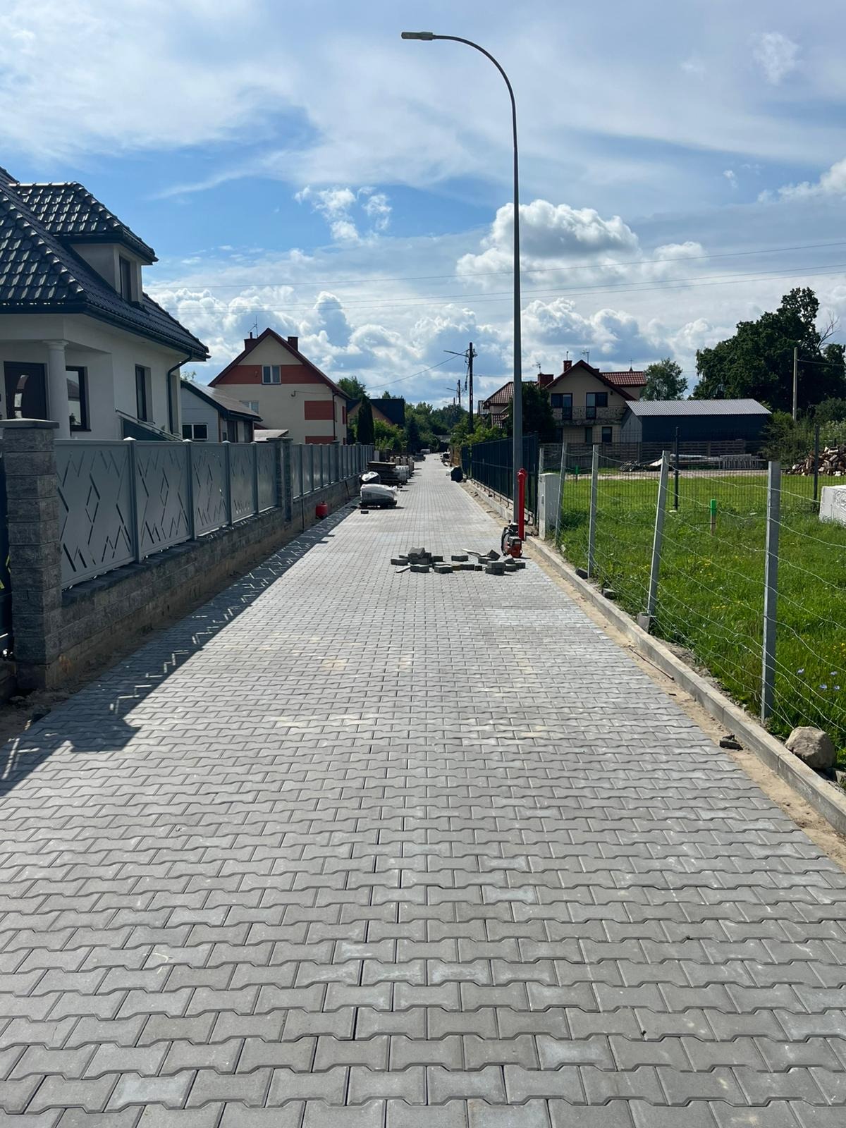 Ułożona szara kostka brukowa na podjeździe, widok w perspektywie, z elementami niezakończonej pracy i domami w tle, w słoneczny dzień.