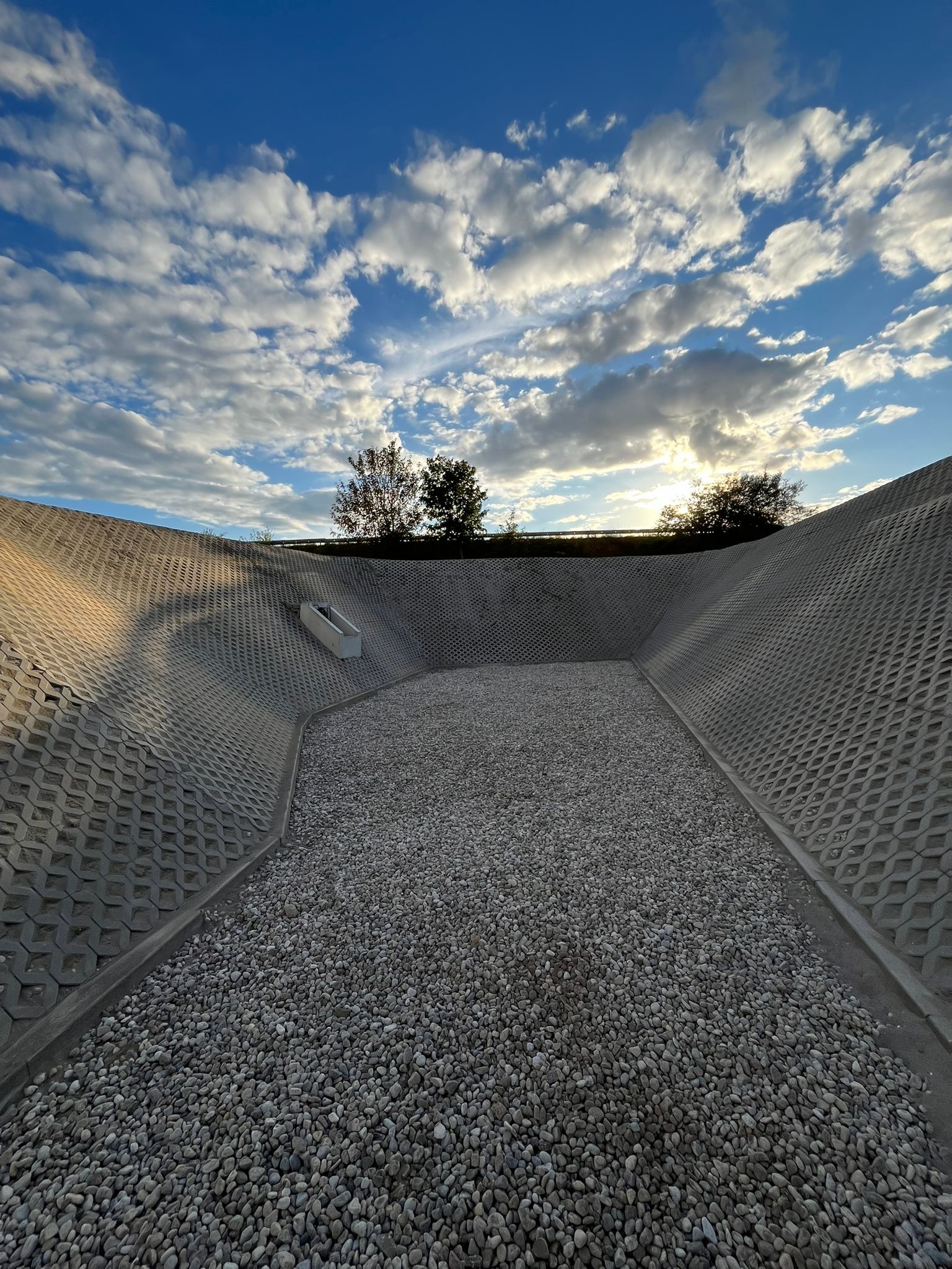 Skarpa umocniona ażurowymi płytami betonowymi, wypełniona żwirem, z widocznym wylotem drenażu i błękitnym niebem w tle. Stabilizacja gruntu.