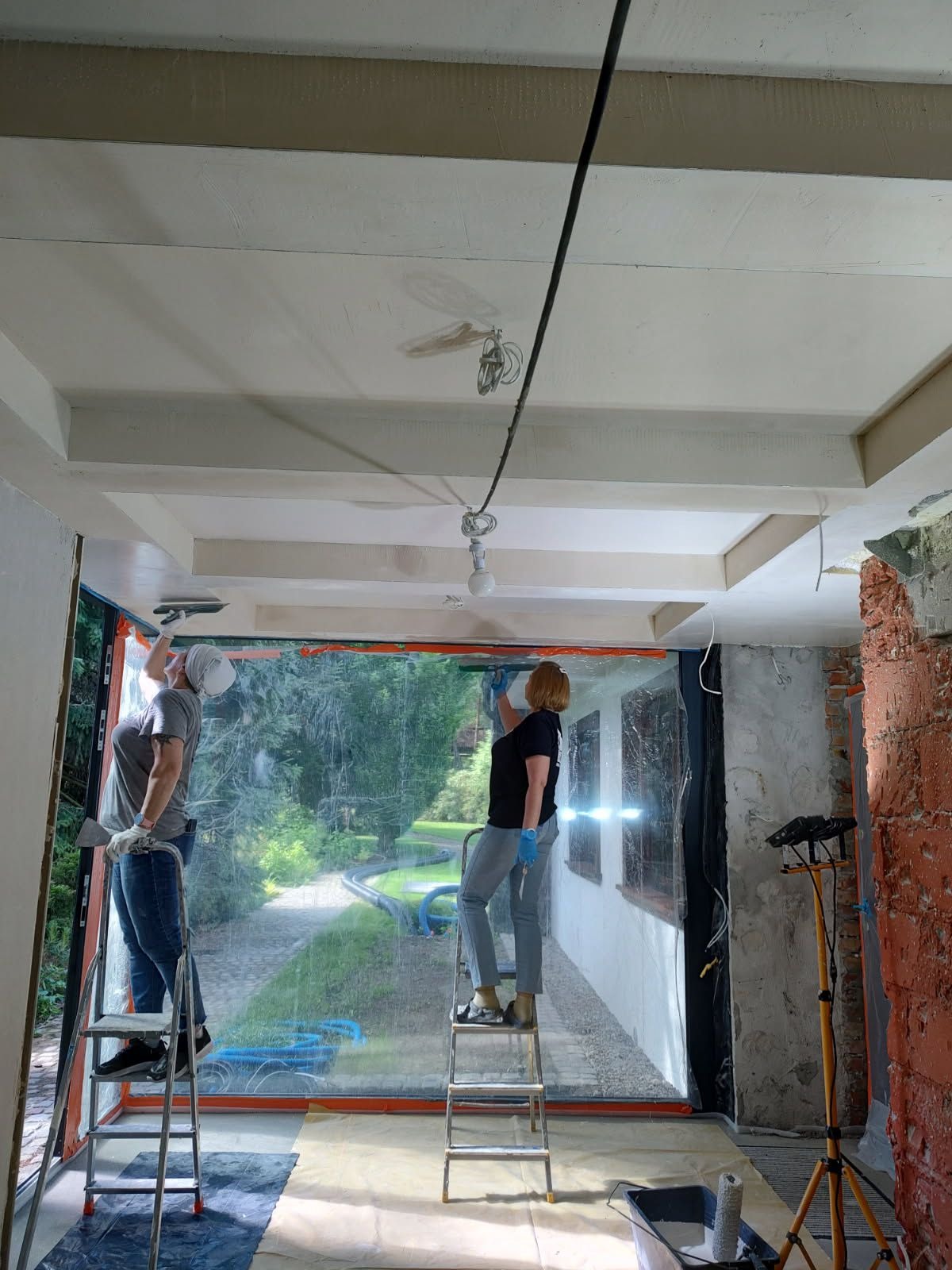 Dwie osoby na drabinach szpachlują sufit w pomieszczeniu z dużym oknem. Widoczne narzędzia, zabezpieczenia i fragmenty surowej ściany z cegły.