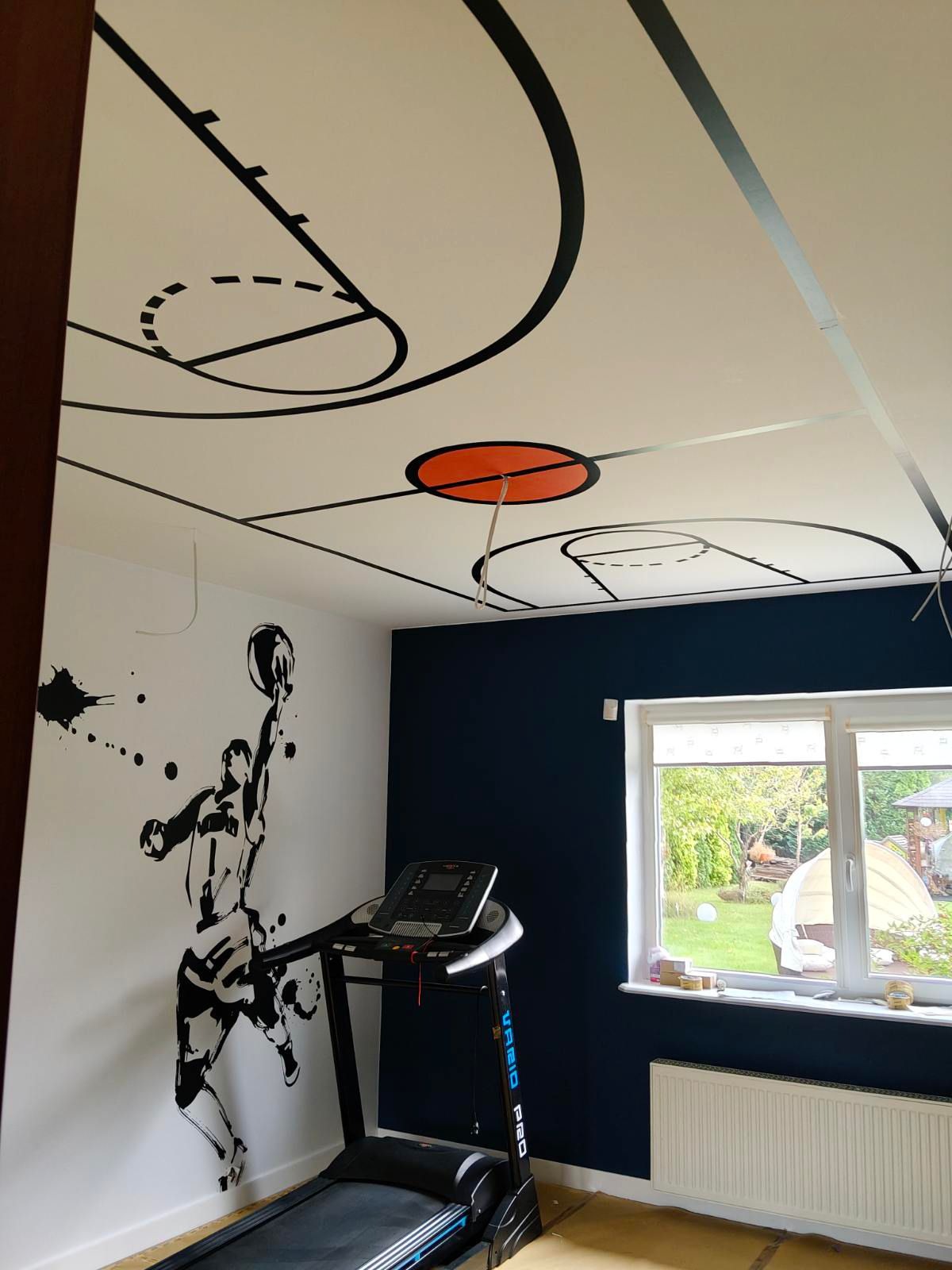 Pokój z motywem koszykówki: malowidło na ścianie przedstawia koszykarza, a sufit imituje boisko. Bieżnia stoi obok, okno z widokiem na ogród w tle.