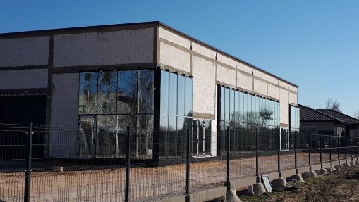 Surowy budynek w trakcie budowy z dużymi oknami, widoczne bloczki betonowe i ogrodzenie z siatki.