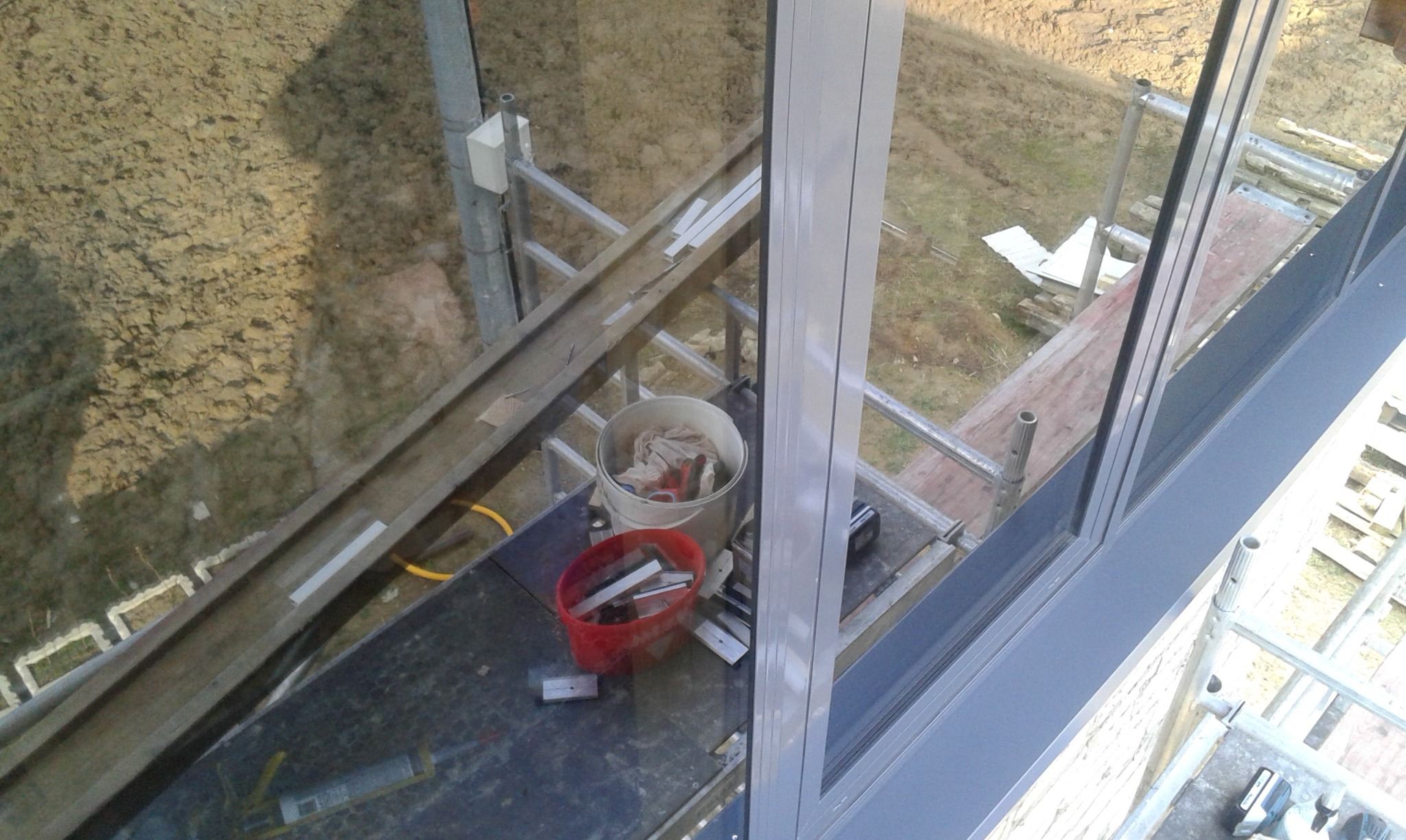 Widok z góry na rusztowanie z wiadrami narzędzi i elementami montażowymi przy oknie, z ziemią w tle.