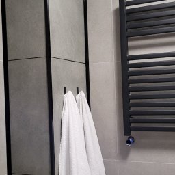 SAMIROL SPÓŁKA Z OGRANICZONĄ ODPOWIEDZIALNOŚCIĄ - Szare płytki w łazience z czarną ramą lustra i grzejnikiem drabinkowym; białe ręczniki wiszą na haczykach.