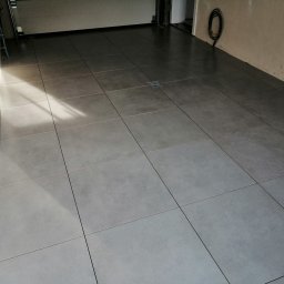 SAMIROL SPÓŁKA Z OGRANICZONĄ ODPOWIEDZIALNOŚCIĄ - Szare, matowe płytki podłogowe ułożone w garażu, widoczny odpływ liniowy z dekoracyjnym wzorem gwiazdy, kabel zwinięty na ścianie i rura kanalizacyjna.