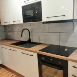 Nowoczesna kuchnia z białymi szafkami, drewnianym blatem, czarną armaturą i płytkami imitującymi beton na ścianie, po remoncie.