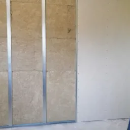 Montaż izolacji z wełny mineralnej w metalowym stelażu pod płytę gipsowo-kartonową, widoczny poziomica oparta o ścianę.