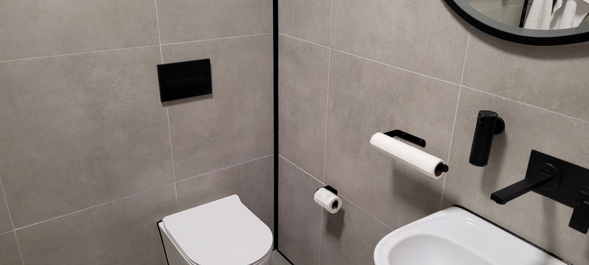 Nowoczesna łazienka wykończona szarymi płytkami, z czarną armaturą, okrągłym lustrem i podwieszaną toaletą.