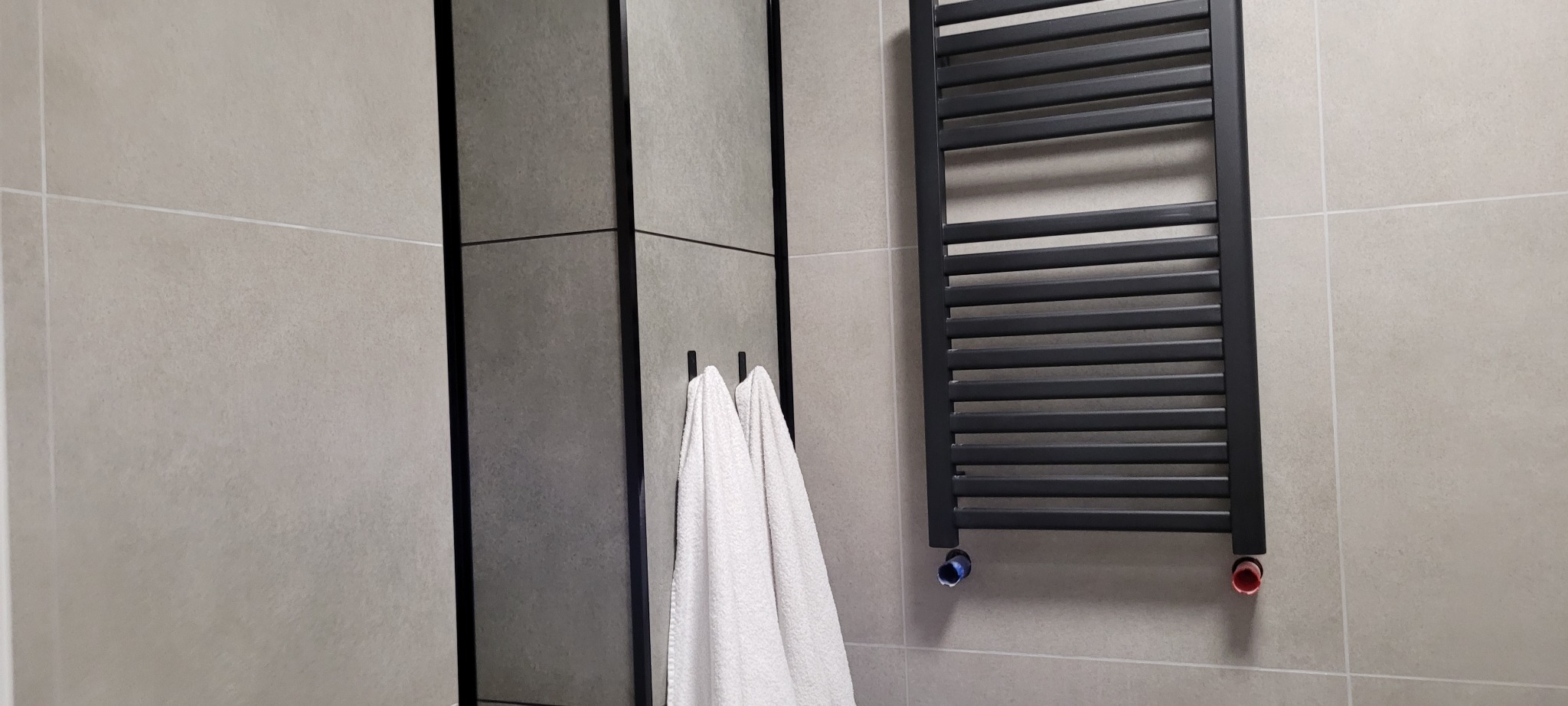 Szare płytki w łazience z czarną ramą lustra i grzejnikiem drabinkowym; białe ręczniki wiszą na haczykach.