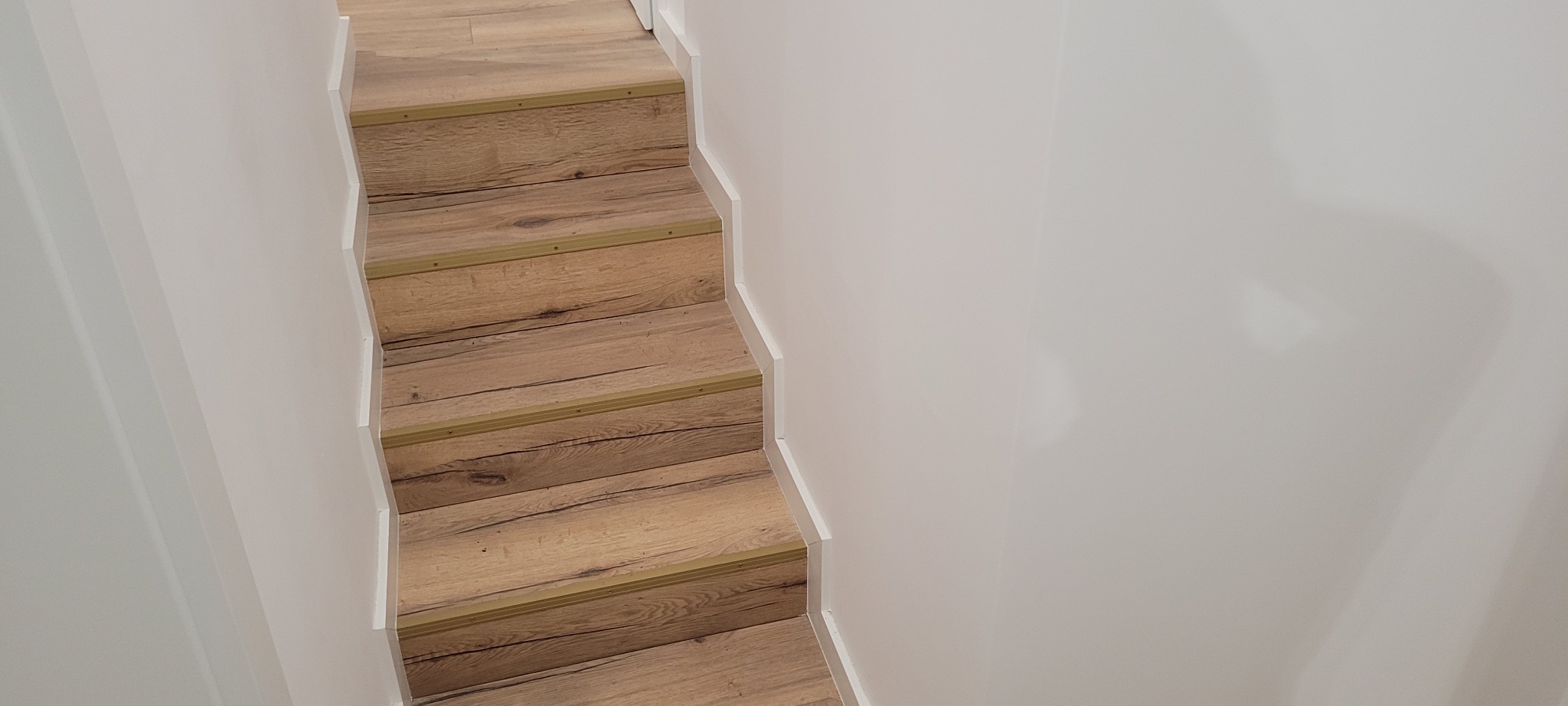 Schody wyłożone panelami drewnopodobnymi z metalowymi listwami antypoślizgowymi, białe ściany i listwy przypodłogowe.