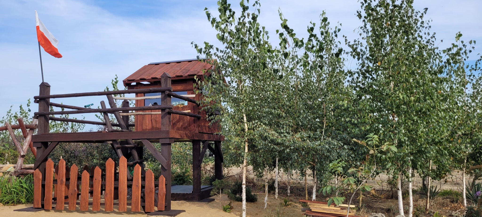 Drewniany domek na drzewie z polską flagą, otoczony brzozami i niskim płotem, w słoneczny dzień.