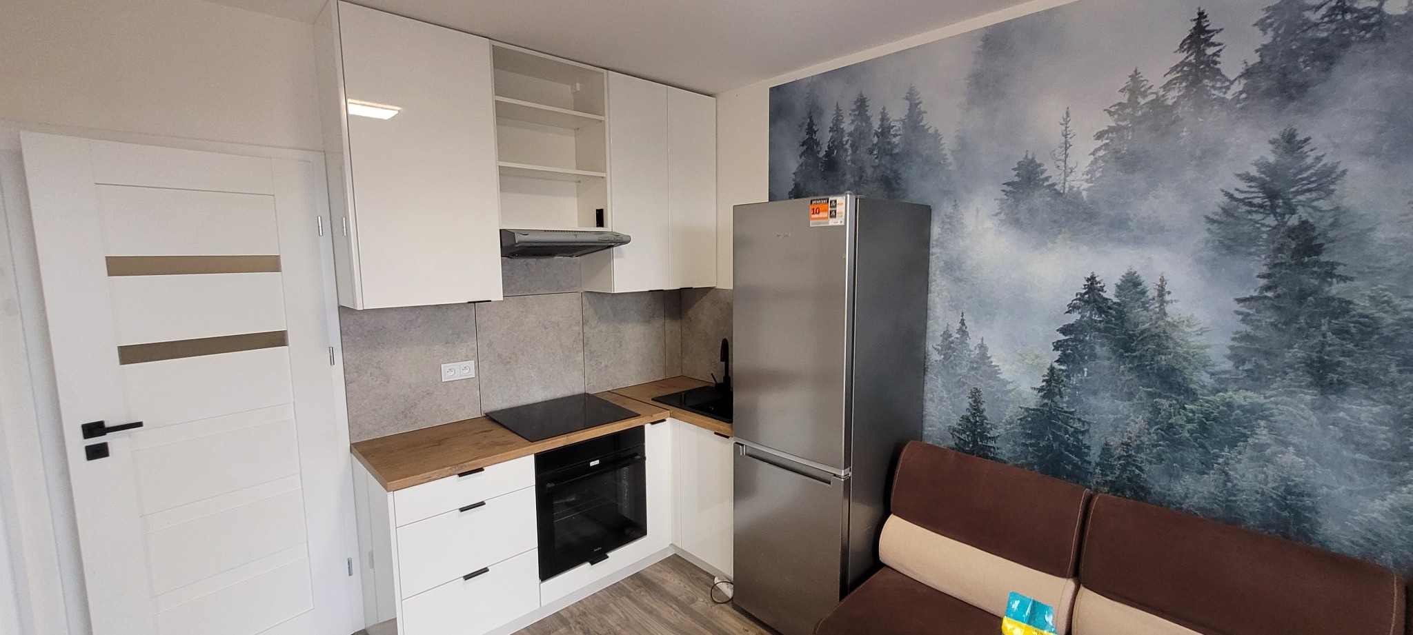 Nowoczesna kuchnia z białymi szafkami, drewnianym blatem i tapetą z motywem leśnym, widziana z perspektywy pokoju dziennego z brązową sofą.