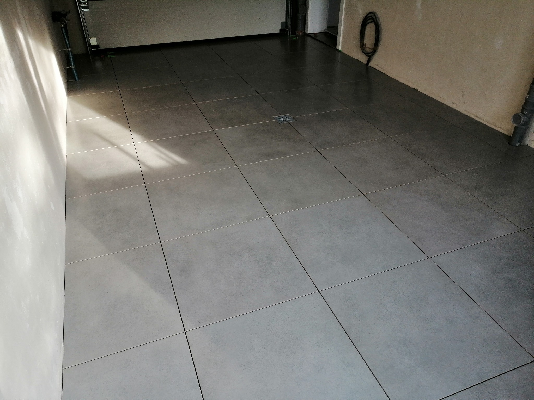 Szare, matowe płytki podłogowe ułożone w garażu, widoczny odpływ liniowy z dekoracyjnym wzorem gwiazdy, kabel zwinięty na ścianie i rura kanalizacyjna.