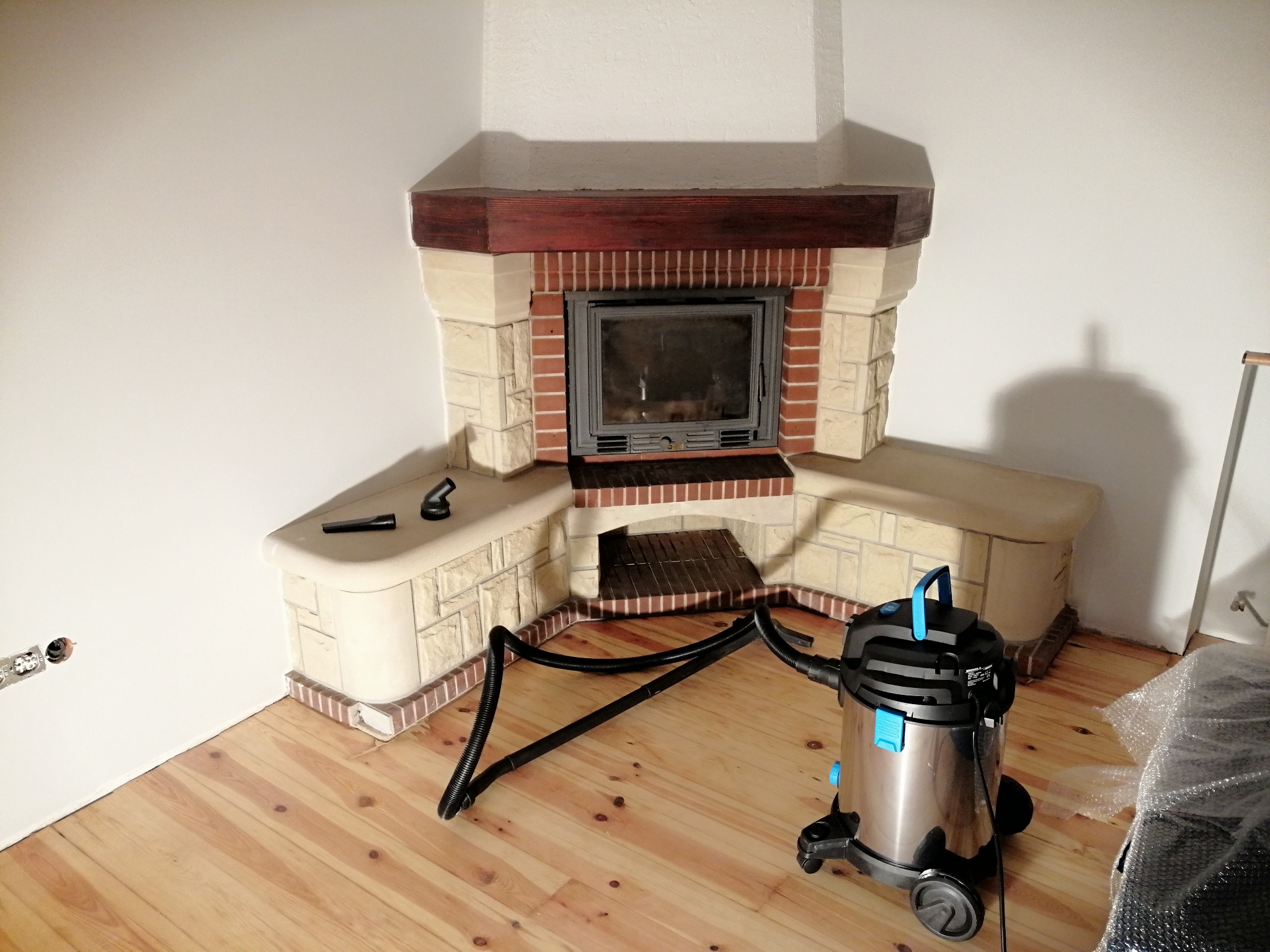 Nowo wykończony kominek z obudową z cegły i kamienia, drewniany gzyms, na podłodze odkurzacz przemysłowy, widoczne gniazdko elektryczne w ścianie i surowe drewniane deski podłogowe.
