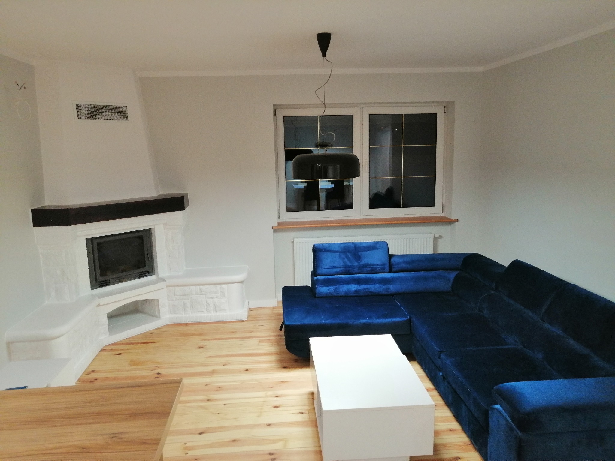 Salon po remoncie: jasne ściany, drewniana podłoga, biały kominek z czarnym wkładem, granatowy narożnik i biały stolik kawowy pod wiszącą czarną lampą.