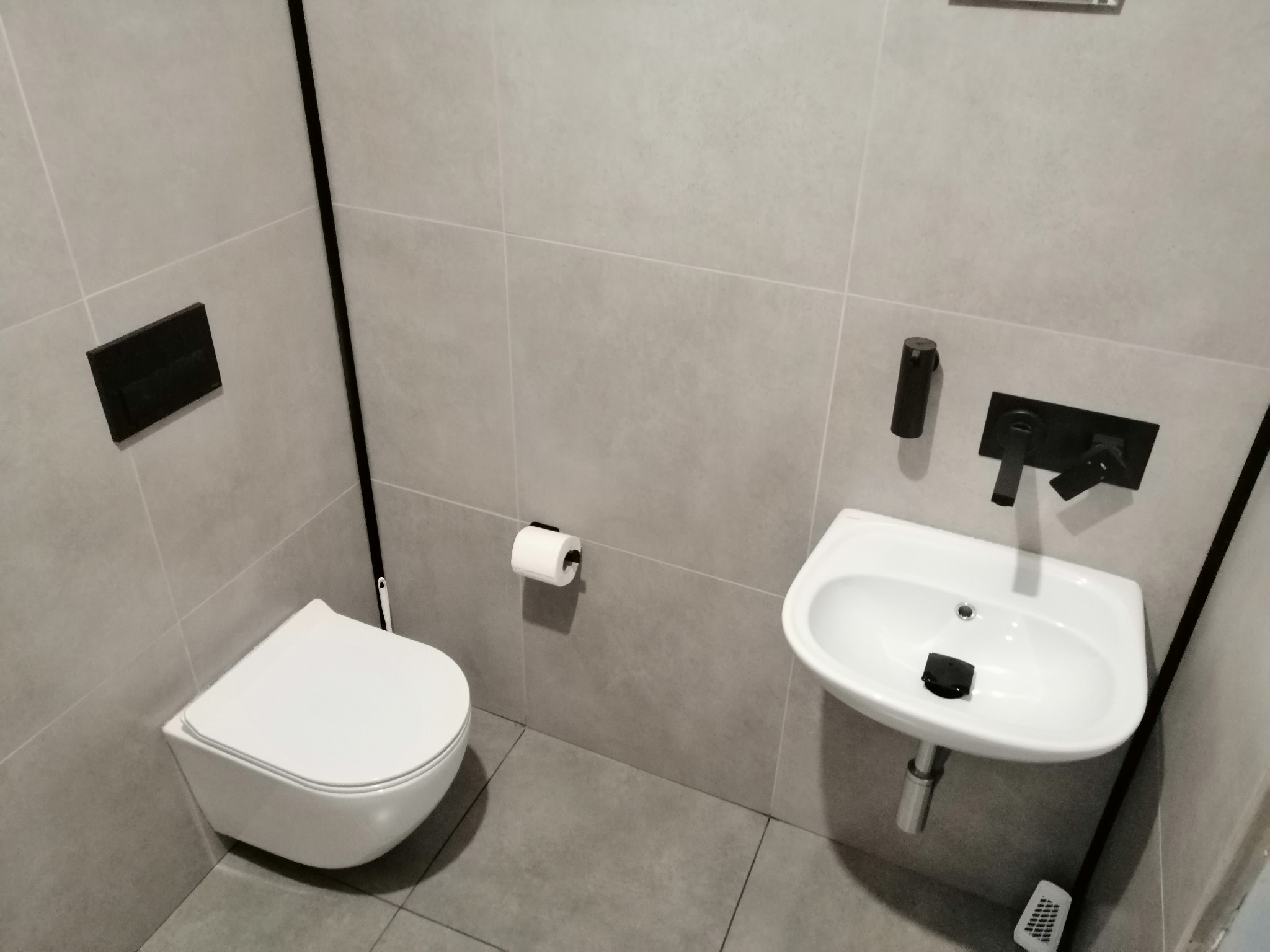 Nowoczesna łazienka z szarymi płytkami, wiszącą toaletą i małą umywalką z czarną armaturą, minimalistyczny design.