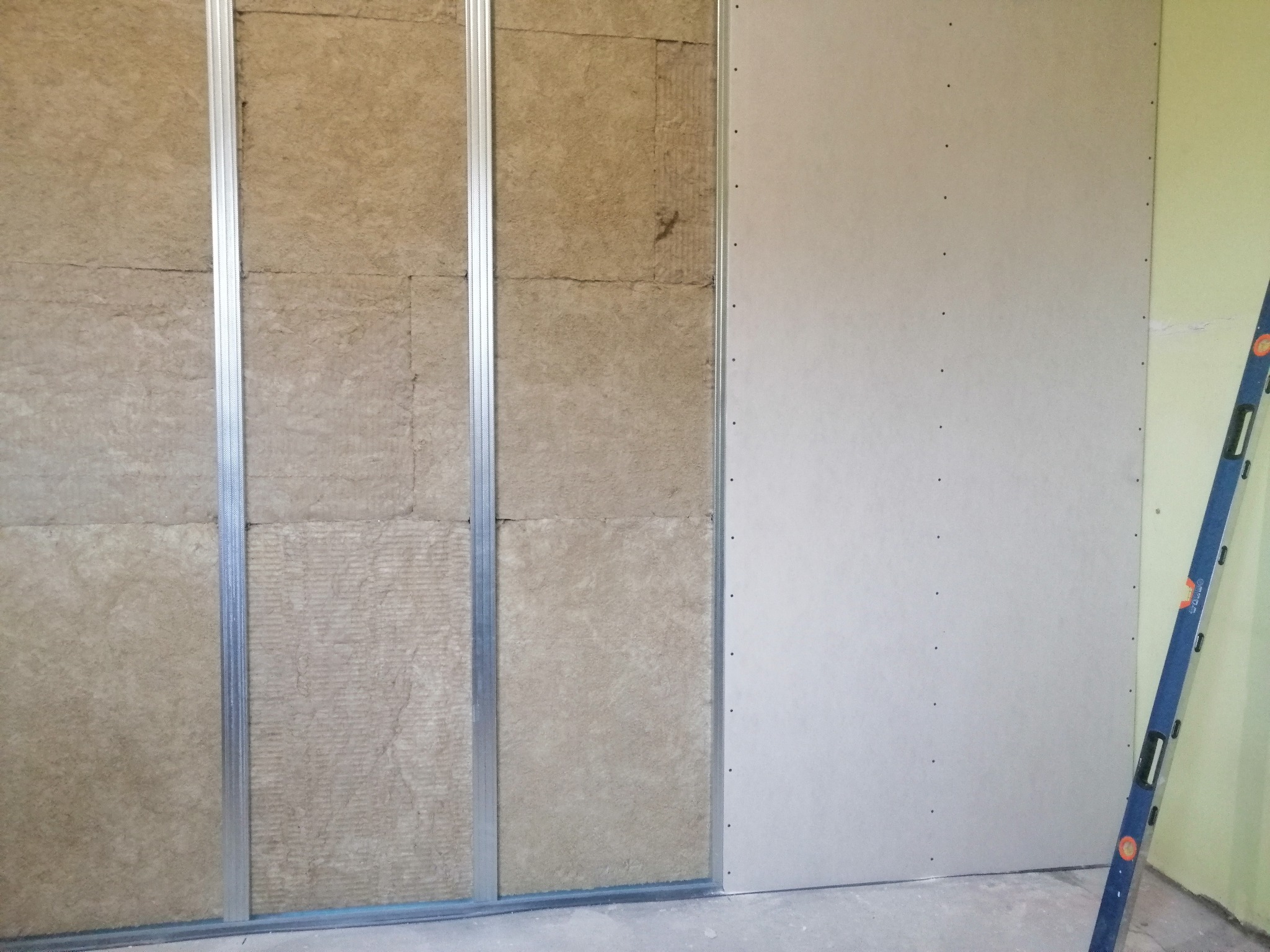 Montaż izolacji z wełny mineralnej w metalowym stelażu pod płytę gipsowo-kartonową, widoczny poziomica oparta o ścianę.