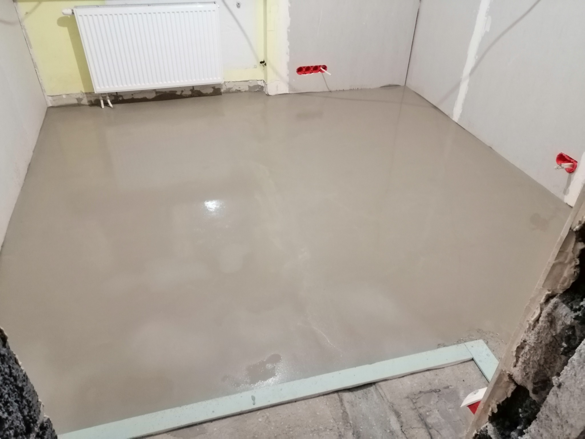 Świeżo wylana, samopoziomująca wylewka podłogowa w trakcie schnięcia, widoczne ślady wilgoci i refleksy światła, ściany przygotowane do dalszych prac, grzejnik na ścianie.