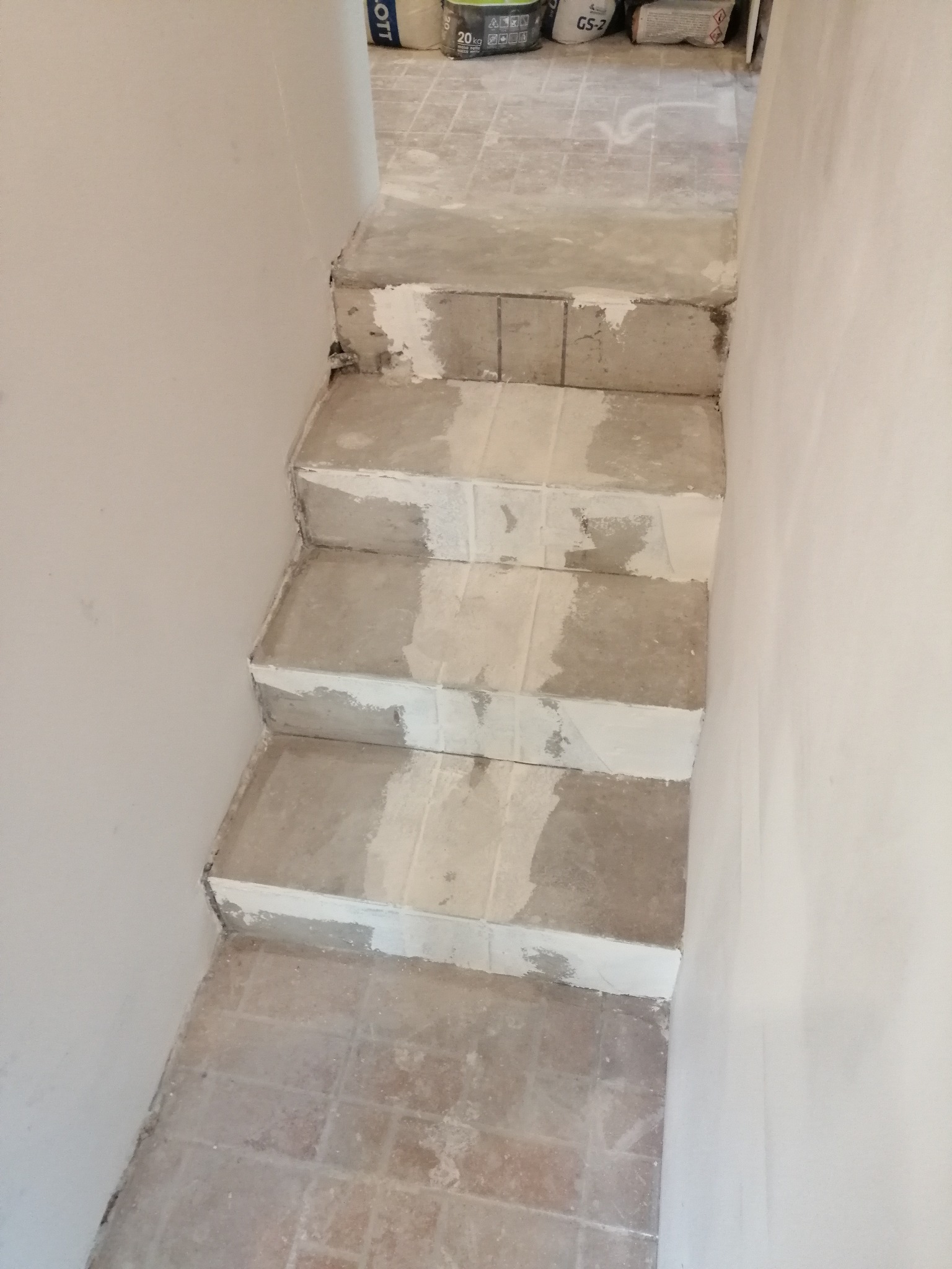 Schody w trakcie prac przygotowawczych do położenia płytek, widoczne ślady zaprawy i kleju, na podłodze resztki materiałów budowlanych, w tle worki z zaprawą.