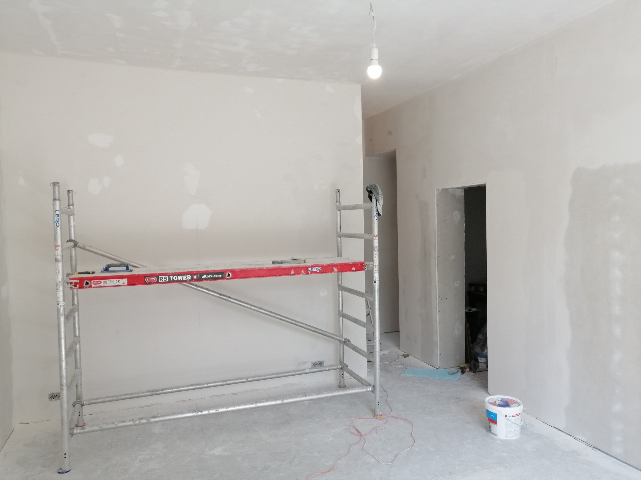 Wnętrze w trakcie szpachlowania: widoczne ściany z nałożonymi łatami gipsu, aluminiowe rusztowanie z czerwonymi elementami, wiadro z farbą i wisząca żarówka