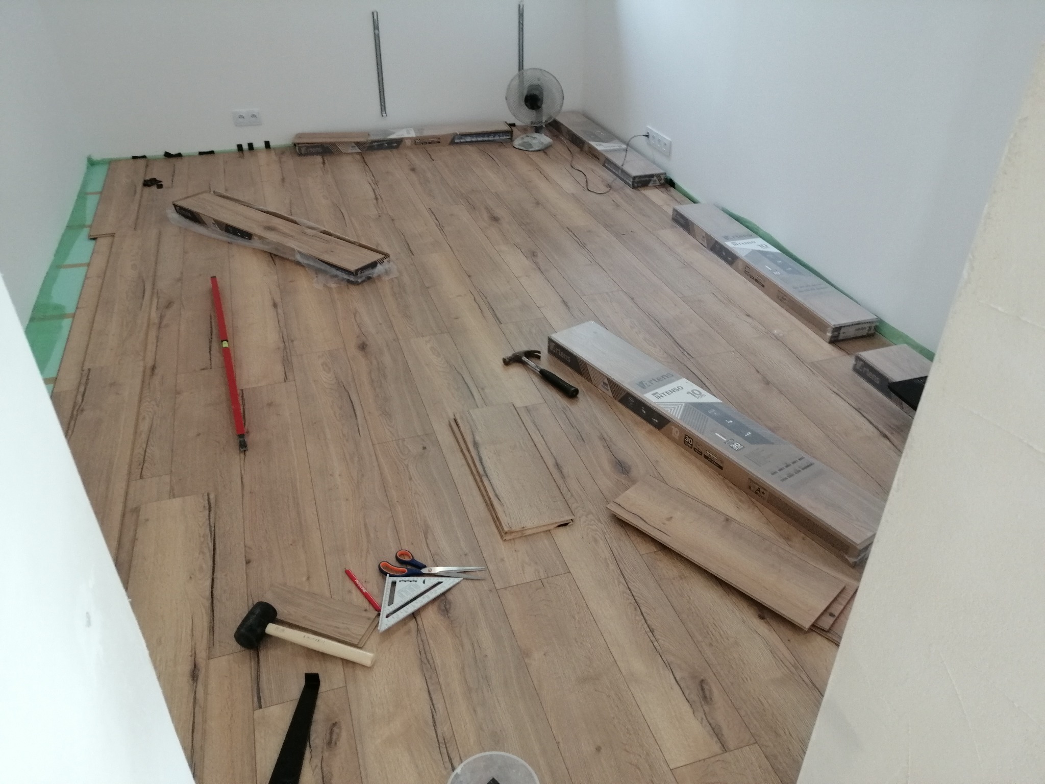 Układanie paneli podłogowych w pokoju: widoczne panele, narzędzia (młotek, kątownik, nożyczki), podkład, wentylator i paczki paneli. Częściowo ułożona podłoga.