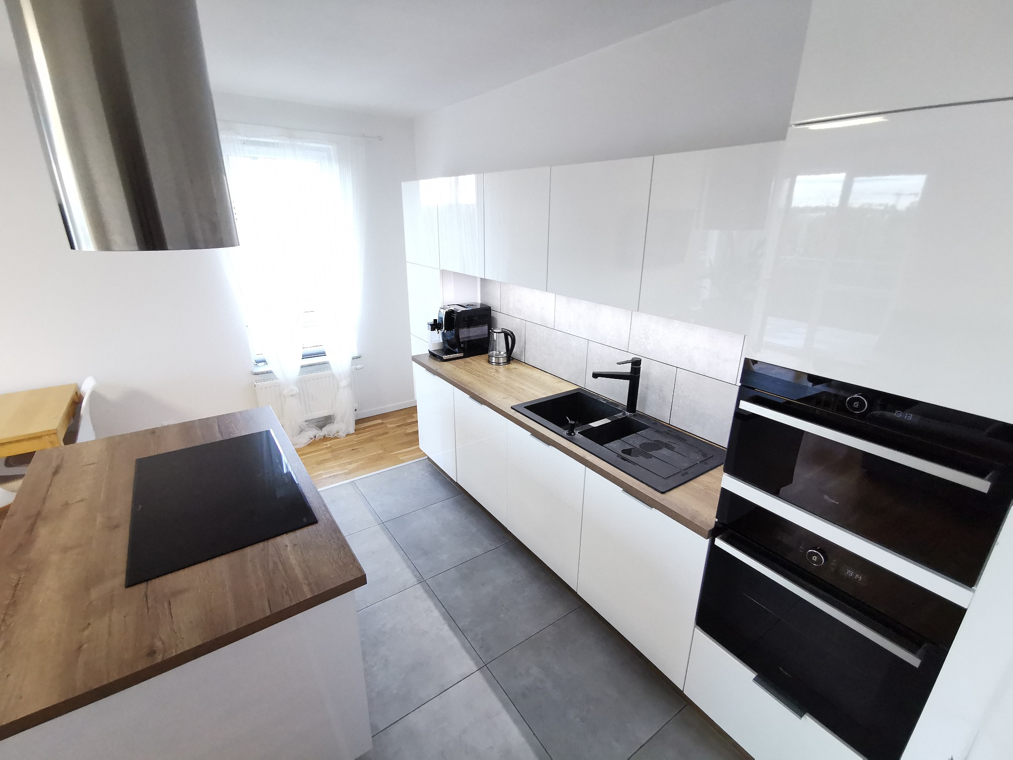 Nowoczesna kuchnia z białymi szafkami, drewnianym blatem i czarną armaturą, widok na wyspę z płytą indukcyjną i okno z białymi zasłonami.