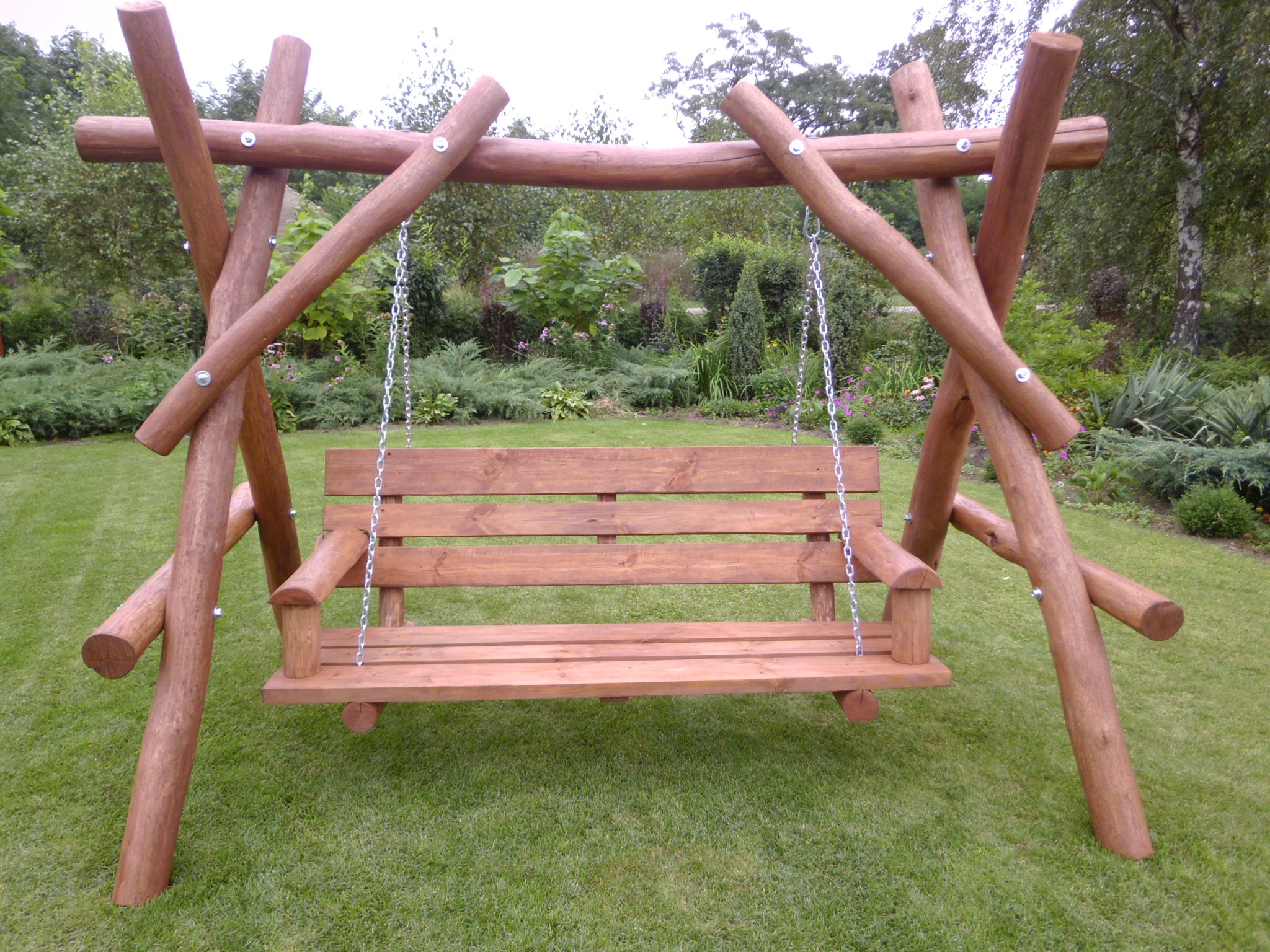 Drewniana huśtawka ogrodowa z bali, zawieszona na łańcuchach, umieszczona na trawniku w zadbanym ogrodzie.
