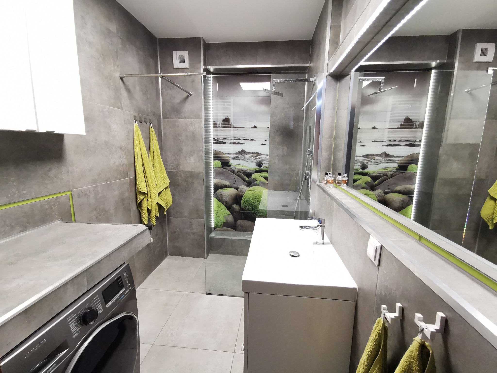 Nowoczesna łazienka z szarymi płytkami, kabiną prysznicową z fototapetą morskiego krajobrazu, białą umywalką i pralką pod blatem.