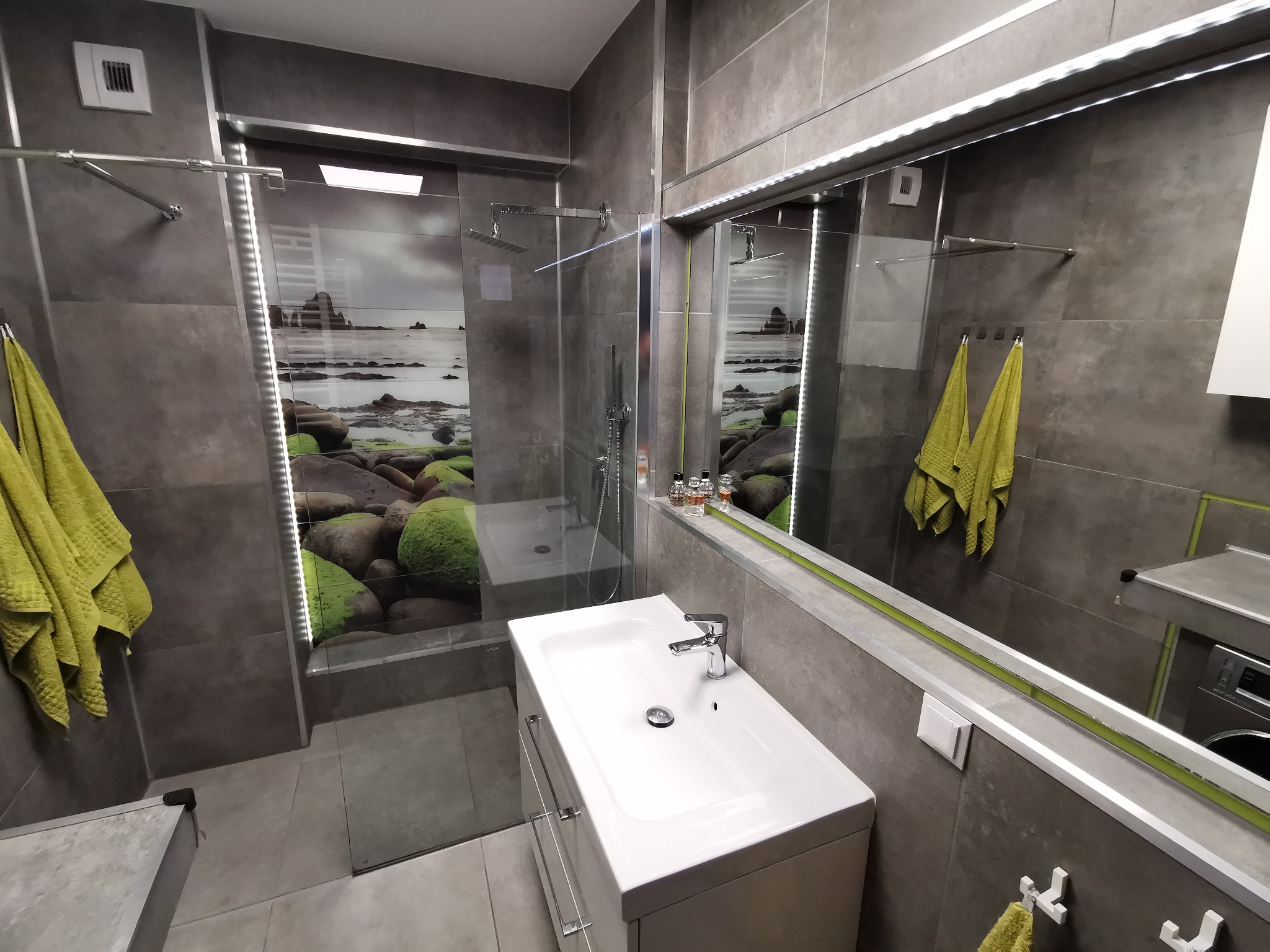 Nowoczesna łazienka z szarymi płytkami, kabiną prysznicową z fototapetą z motywem morskim, białą umywalką nablatową i zielonymi akcentami w postaci ręczników i listwy przy lustrze.