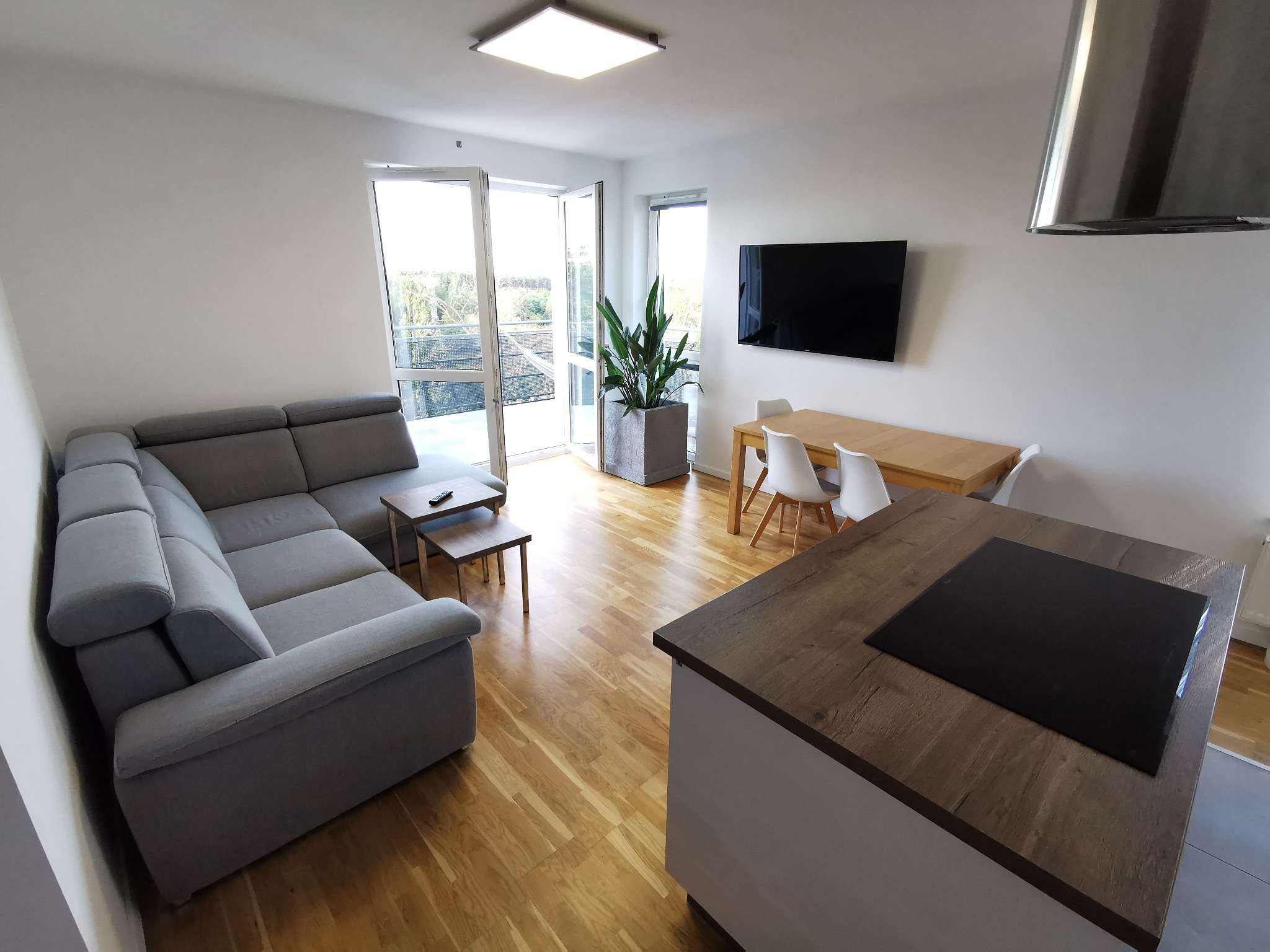 Jasne, nowocześnie urządzone wnętrze mieszkania z szarym narożnikiem, drewnianym stołem jadalnym i wyspą kuchenną z płytą indukcyjną, podkreślone parkietem w ciepłym odcieniu drewna.