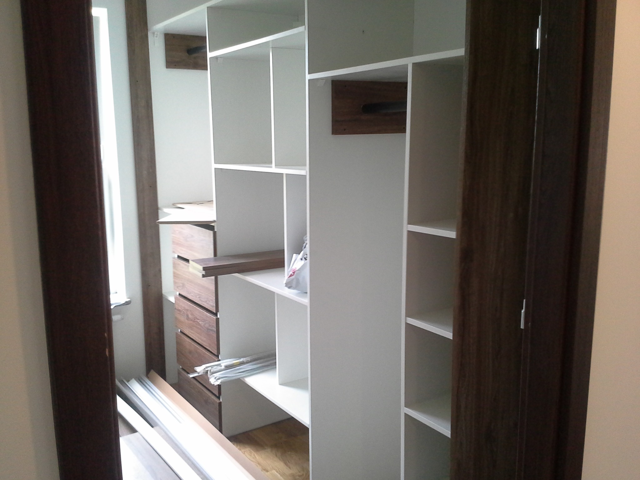 Wnętrze szafy wnękowej w trakcie montażu, widoczne białe półki, drewniane szuflady oraz elementy wykończeniowe w kolorze ciemnego drewna, częściowo zamontowane, z elementami do dalszego montażu...