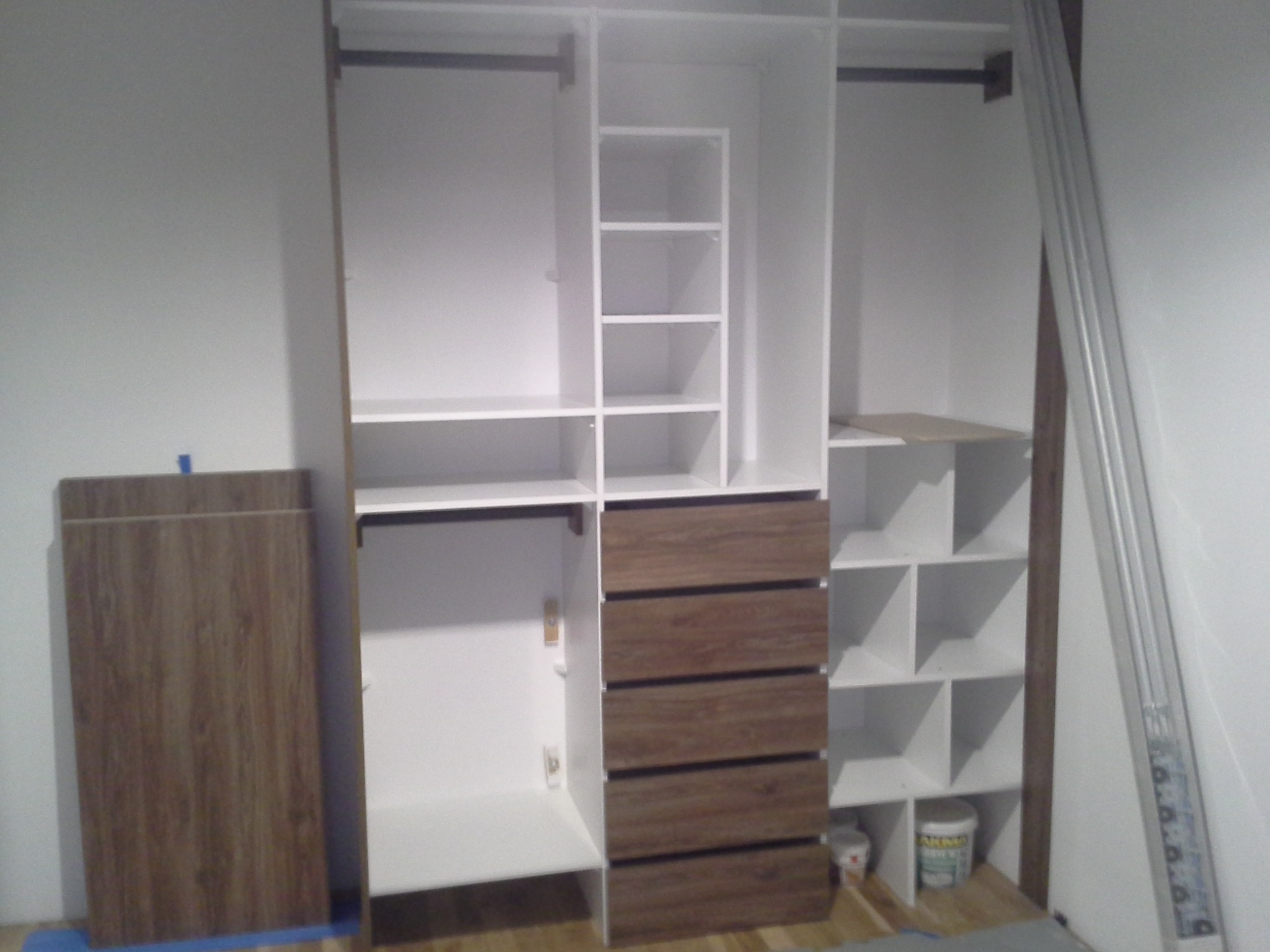 Wnętrze szafy wnękowej w trakcie montażu, widoczne białe półki, brązowe szuflady, płyty laminowane oparte o ścianę, puszki z farbą w dolnej części.