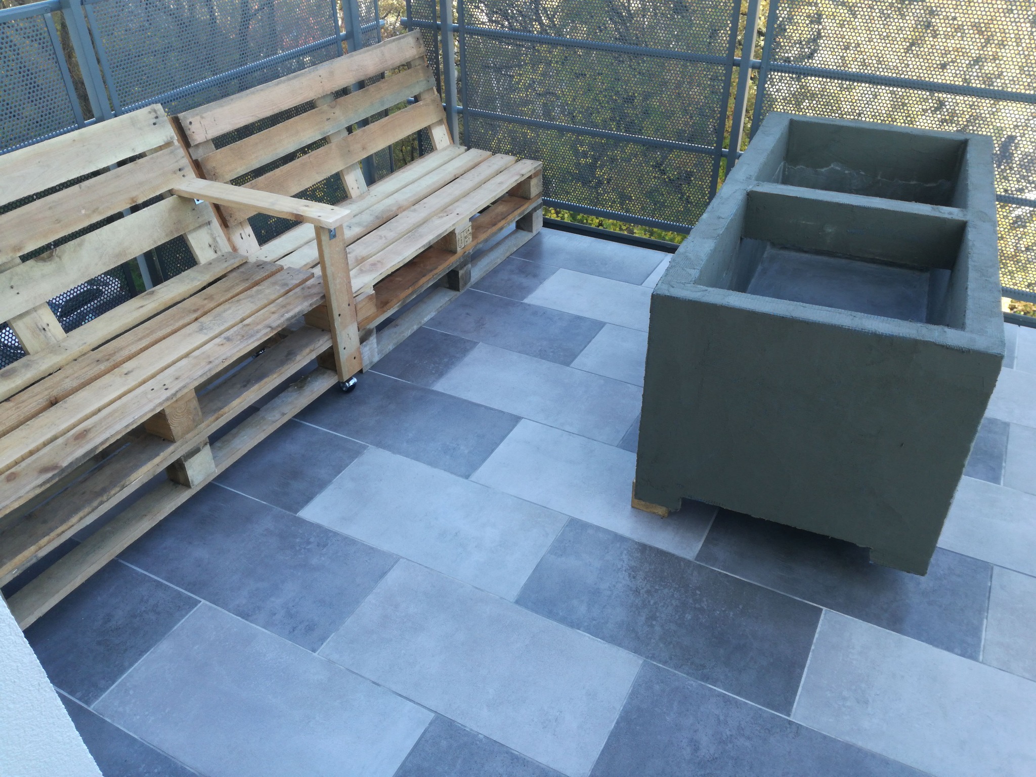 Balkon z szarymi płytkami, ławką z palet i betonową donicą z dwiema komorami.